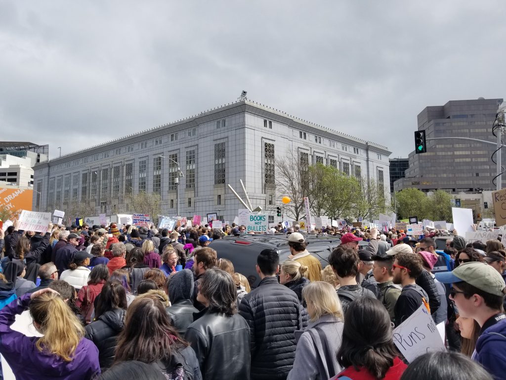 Protesters gather at the March for Our Lives in San Francisco, holding signs that say "Books not Bullets," "Puns not Guns," and "I shouldn't have to die for my education."