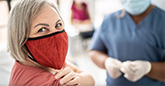older woman with red mask pulling up her sleeve in preparation for a COVID-19 vaccine. Practitioner of color wearing scrubs stands in the background.