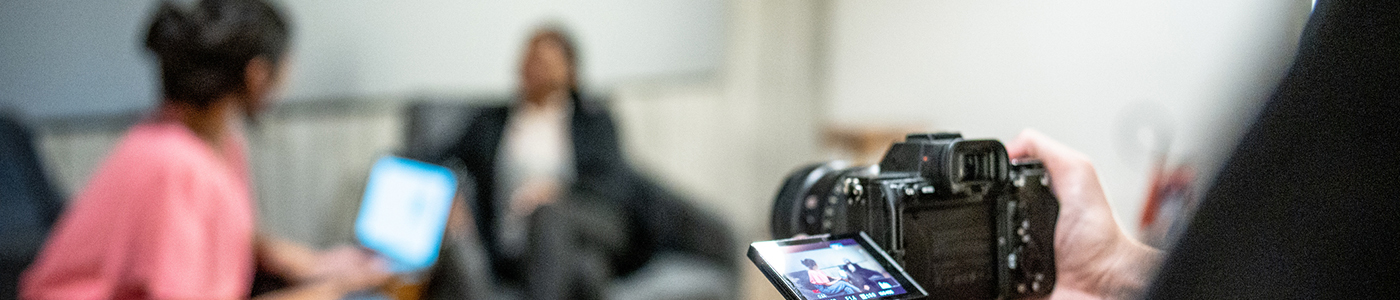 A journalist interviews a source while both are seated on a couch. A photographer captures the session from the opposite side of the room.