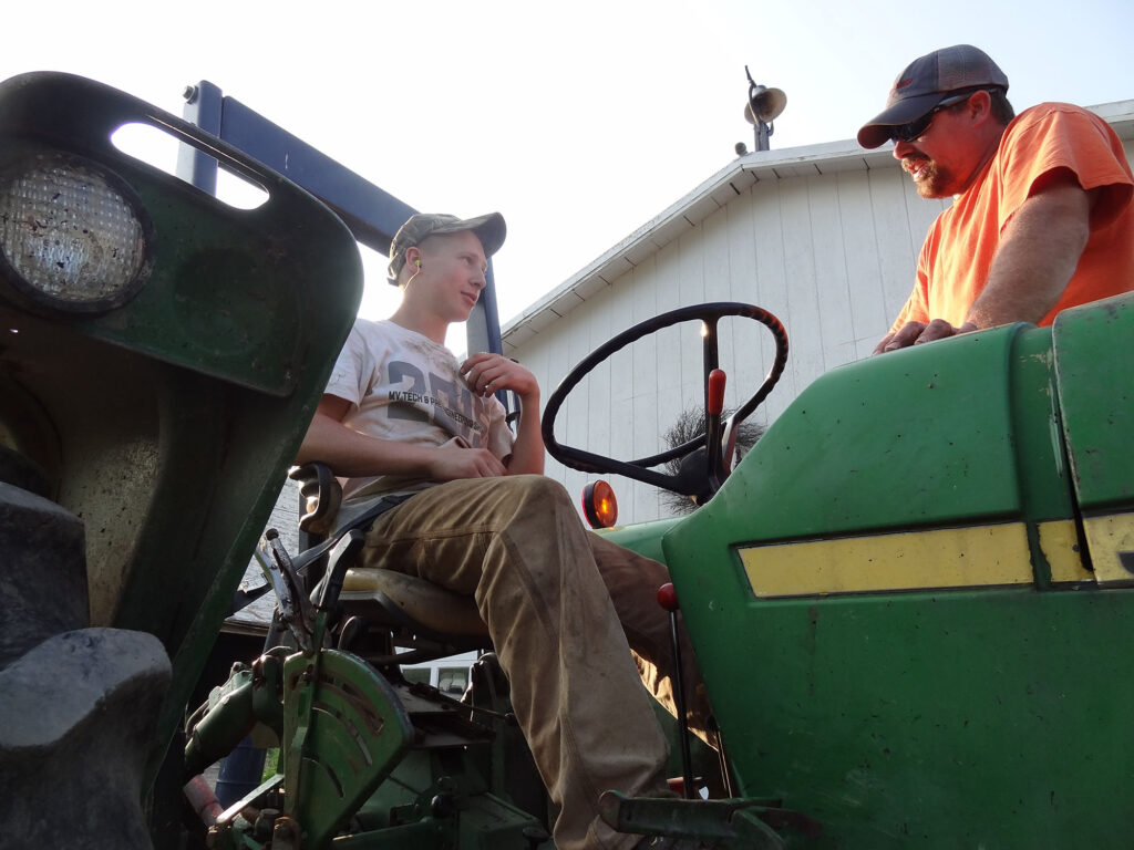 Two farmers speak to each other. One is seated on a tractor, the other standing and leaning in. 